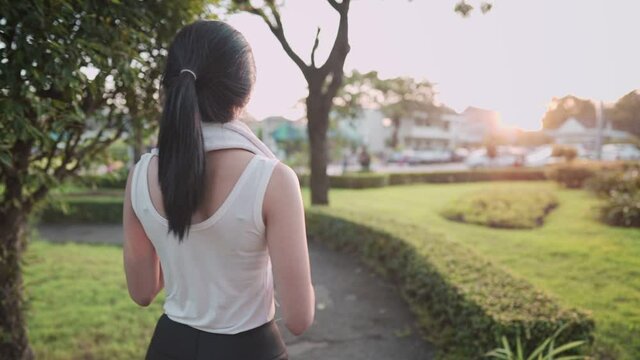 Back view follow up: young athletic healthy skin woman walks on run way to cold down after work out and wipes out sweat using white towel on her neck in green beautiful park, healthy and lifestyle 
