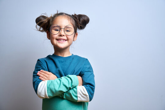 Naklejki Portrait of cute swarthy caucasian little girl with glasses, arms crossed and funny hairstyle, beautiful happy child smiling and laughing looking at camera isolated in studio