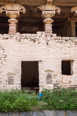 Fototapeta premium Hampi, Karnataka, India - November 4, 2013: Hampi Bazar street. Historic ruinous empty bulding with decorated roof holding pillars and frescos on white painted wall. Green foliage in front.