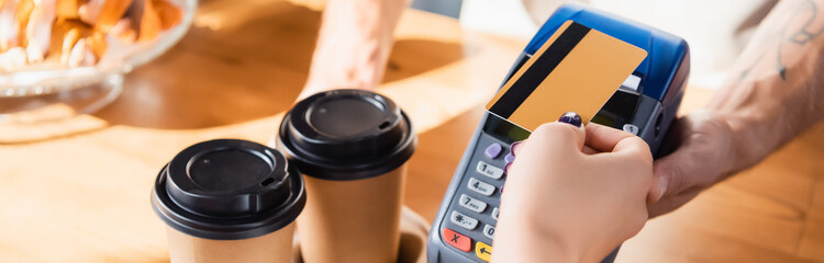 cropped of woman paying with credit card through terminal near takeaway coffee in cafe, banner