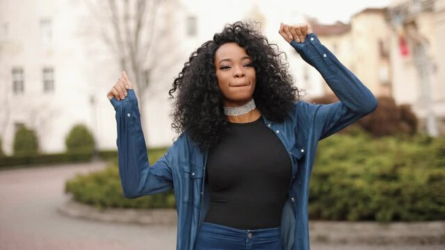 Close-up portrait of the young attractive african girl with natural make-up and pretty smile actively dancing and shaking her dark curly hair outdoor.