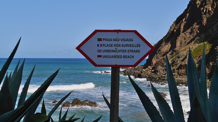 Metal sign above rocky beach Praia da Maiata between agave leaves in village Porto da Cruz in the north of Madeira island, Portugal informing that the beach is unguarded in different languages.
