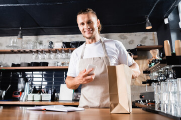 Fototapeta na wymiar barista in apron holding mobile phone near notebook and paper bag on bar counter