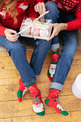 Unfold Christmas presents. A young guy and a girl are sitting on the floor in sweaters with deer and funny Christmas socks. Untie the ribbon on the marshmallow box. Receive gifts in the new year.