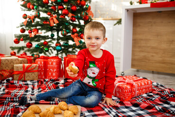 The boy who sits near the Christmas tree for the new year. Christmas decor with gifts, a child near the Christmas tree eats croissants and smiles