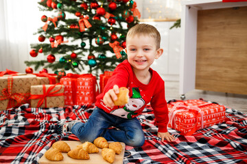The boy who sits near the Christmas tree for the new year. Christmas decor with gifts, a child near the Christmas tree eats croissants and smiles