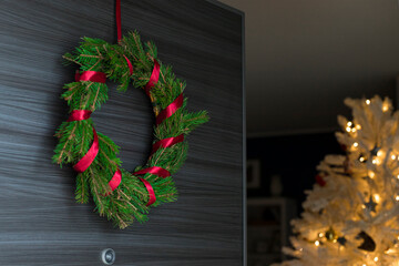 Foraged diy pine wreath with red ribbon on door opening with white, illuminated christmas tree...