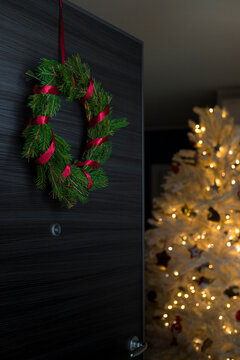 Foraged diy pine wreath with red ribbon on door opening with white, illuminated christmas tree inside the house. Vertical shot.