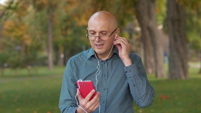 Slow Motion Bald senior man in glasses sits on the bench in park and communicates via smartphone