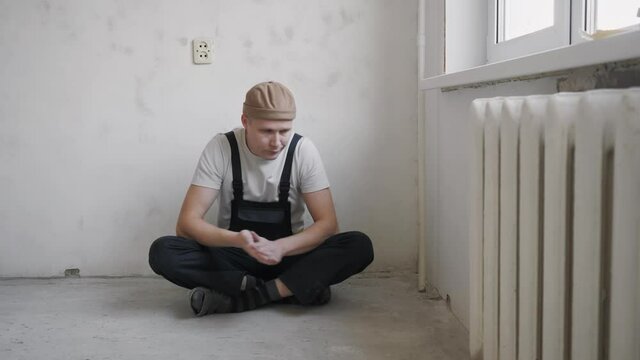A man in construction overalls is resting sitting on the floor near the wall after the work done to repair the apartment. High quality 4k footage