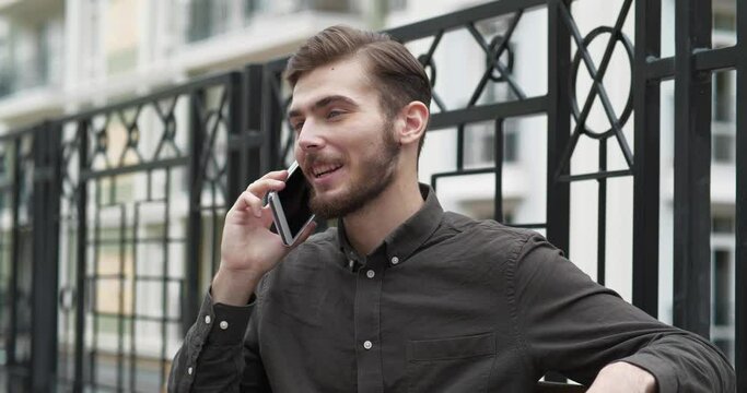 Young man sits on the bench and talks on smartphone, he smiles and he is in a good mood