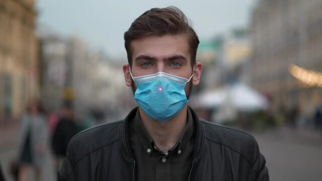 Young man in medical protective mask stands on the street and looks at the camera. Red laser beam of the sight rises and shines on the forehead, man closes his eyes and waits shot