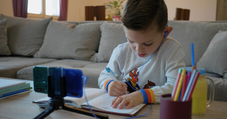 A boy at home follows school classes on his phone and writes in a notebook. Children have virtual school. On the screen homeschooling