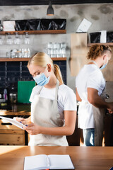  blonde barista in medical mask using digital tablet near colleague on blurred background