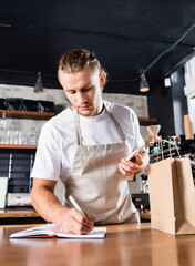 barista holding mobile phone and writing in notebook near paper bag on bar counter