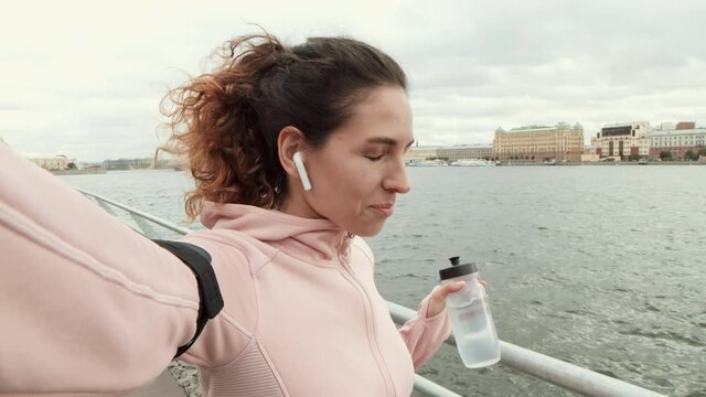 Medium shot of attractive Caucasian sportswoman wearing wireless headphones standing on embankment in city center after jogging, shooting herself on video, drinking water and laughing