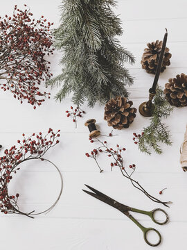 Making christmas rustic wreath, red berries, pine cones, scissors on white wooden table, top view