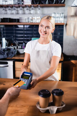  blonde barista holding payment terminal near man with credit card and disposable cups on bar counter