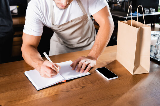 Cropped Of Barista Writing In Notebook Near Paper Bag And Smartphone With Blank Screen On Bar Counter