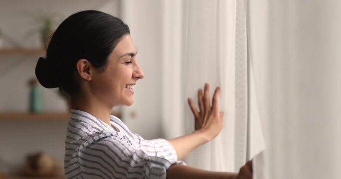 Close up side profile view Indian attractive woman opens curtains in own apartment in morning. Young pretty female enjoy weekend alone, city view through window feels carefree. New happy day concept