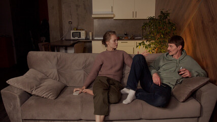 Obraz premium A typical family evening, a guy invites a girl to sit with him on the couch and drink wine. They have smartphones in their hands.