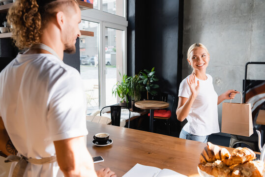  blonde woman holding paper bag and waiving hand to barista on blurred foreground