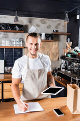 Fototapeta na wymiar , handsome barista looking at camera while writing in notebook and holding digital tablet with blank screen