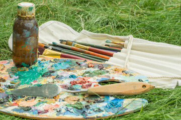 Art supplies for painting with oil paints on green grass. A palette with mixed oil paints, a palette knife, brushes in a case, a bottle of linseed oil. Soft focus