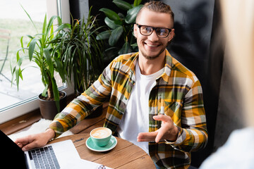  freelancer in eyeglasses and plaid shirt pointing with hand near coffee cup and laptop in cafe