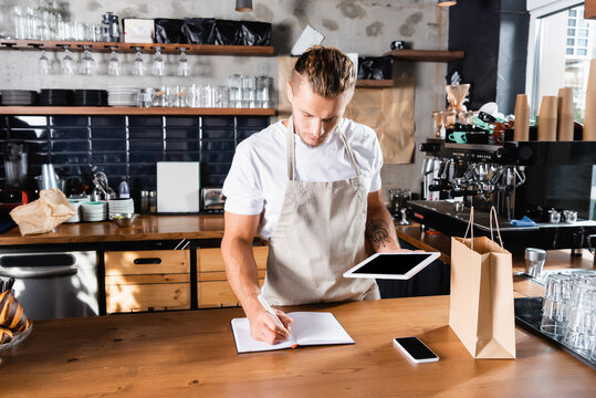  waiter writing in notebook while holding digital tablet near smartphone and paper bag