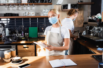 Fototapeta na wymiar waiter in medical mask holding digital tablet near notebook, coffee to go, and barista working on background