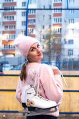 Beautiful woman weared in pink sweater and hat with ice skates on the back walks in winter snowy...