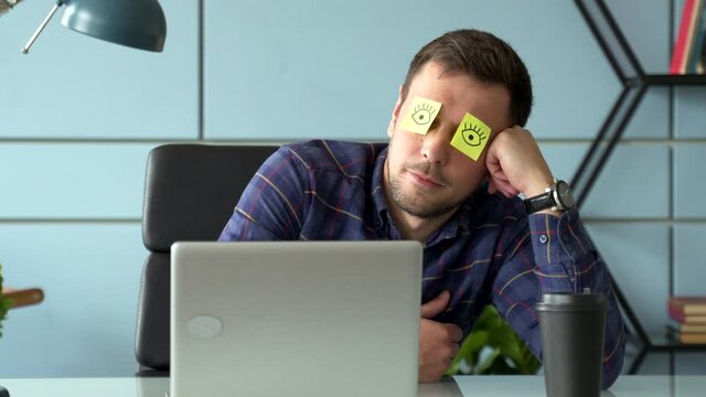 Tired comic male manager pretends working sleeping with stickers on face sits at desk with laptop. 