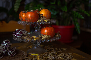 Fruit holder on wooden table with persimmon and red radicchio with a base of dry yellow leaves. Green plants background