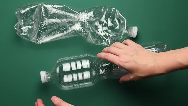 POV female hands crumple and crush transparent PET plastic bottle for recycling. Crumpled used bottles on green background. First point view preparation of recycled household trash as zero waste