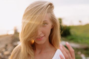 Close up portrait of pretty woman with blond hair posing at camera in sunlight on the shore of ocean with green plants