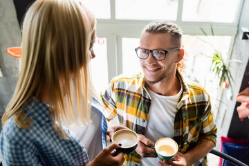  young man in eyeglasses and woman on blurred foreground holding cups of coffee in cafe