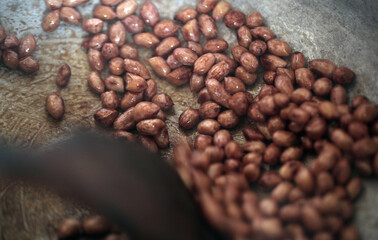horizontal closeup photography of fresh soaked peanuts being roasted on a silver iron pot, with a wooden ladle, indoors in the Gambia, Africa