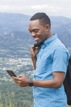 side view of one black skinned young hispanic man outdoor standing happy smiling looking to his smart phone with nature park in background in in dominican republic