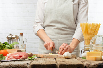The chef cuts with knife garlic on wooden board for cooking pasta alla carbonara. Backstage of preparing traditional italian dish on white background. Frozen motion. Cookbook illustration.