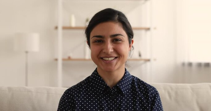 Attractive Indian woman sit on couch looking at camera wave hand start videocall conversation with family using video conference app and device. Video call event, easy comfort chat distantly concept