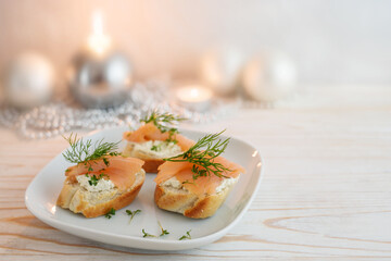 Festive canapes from baguette bread with smoked salmon, cream cheese and dill garnish on a plate, white painted wooden table with candles and Christmas decoration, copy space