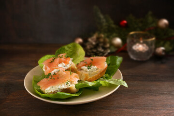 Festive canapes from baguette bread with salmon and cream cheese on green lettuce on a dark rustic wooden table with Christmas decorations, copy space, selected focu
