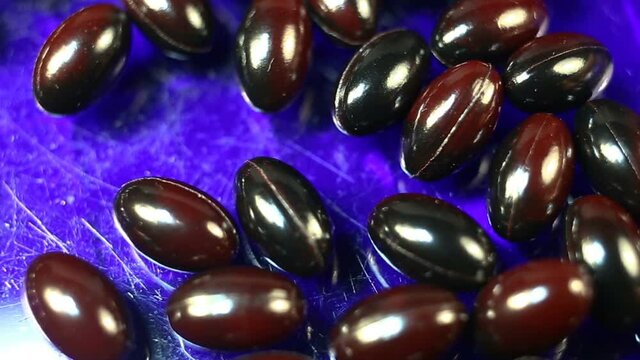 Maroon and black oval spindle-shaped capsules in a shiny gel shell move smoothly from bottom to top. Real time, natural light, on a blue background, carotenoids, carotenoid pigments, Astaxanthin
