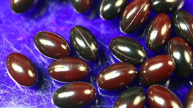 Maroon and black oval spindle-shaped capsules in a shiny gel shell move smoothly from right to left. Real time, natural light, on a blue background, carotenoids, carotenoid pigments, Astaxanthin