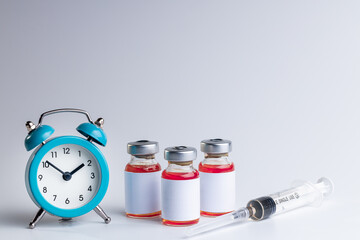medical vaccine vials with red liquid and alarm clock over gray background. time to vaccination...