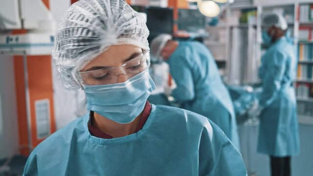Portrait of the young female caucasian assistant or intern wearing medical face mask standing at the medical ward with doctors and preparing for the operation. Patient on the operating table