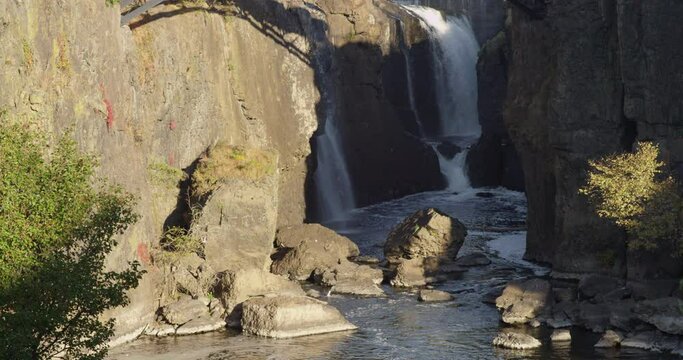 A huge waterfall in a city park with a footbridge over the cliffs during sunny autumn morning, slow motion