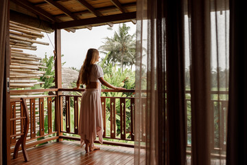 Back view on a pretty  young woman on vacation, standing on the balcony and looking outside on palm trees and tropical jungle