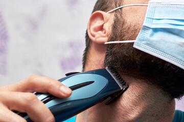 A bearded man with a medical mask on his face tries to shave with a trimmer.